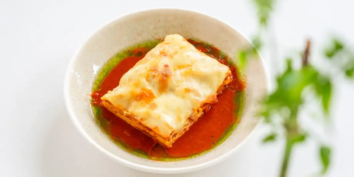 Auf einem weißen Teller liegt ein Stück überbackene Lasagne in roter Tomatensauce, umgeben von etwas grünem Öl, vor einem hellen Hintergrund mit unscharfen Pflanzen.