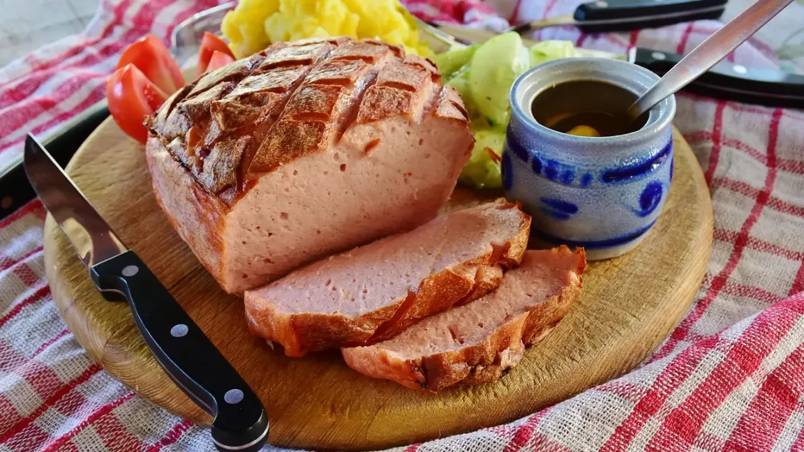 Auf einem Holzbrett liegen ein aufgeschnittener Leberkäse-Laib mit brauner Kruste und mehrere dicke Scheiben, dazu Kartoffelpüree, Tomaten, Salat und ein Schälchen mit Soße; daneben liegt ein Messer auf einem karierten Tischtuch.