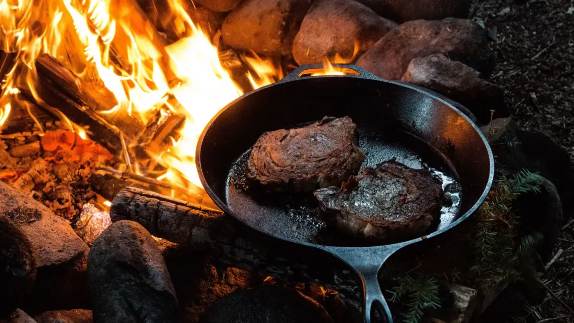 Eine gusseiserne Pfanne mit zwei angebratenen Steaks steht über einem offenen Holzfeuer im Freien, umgeben von Brennholz und Steinen.