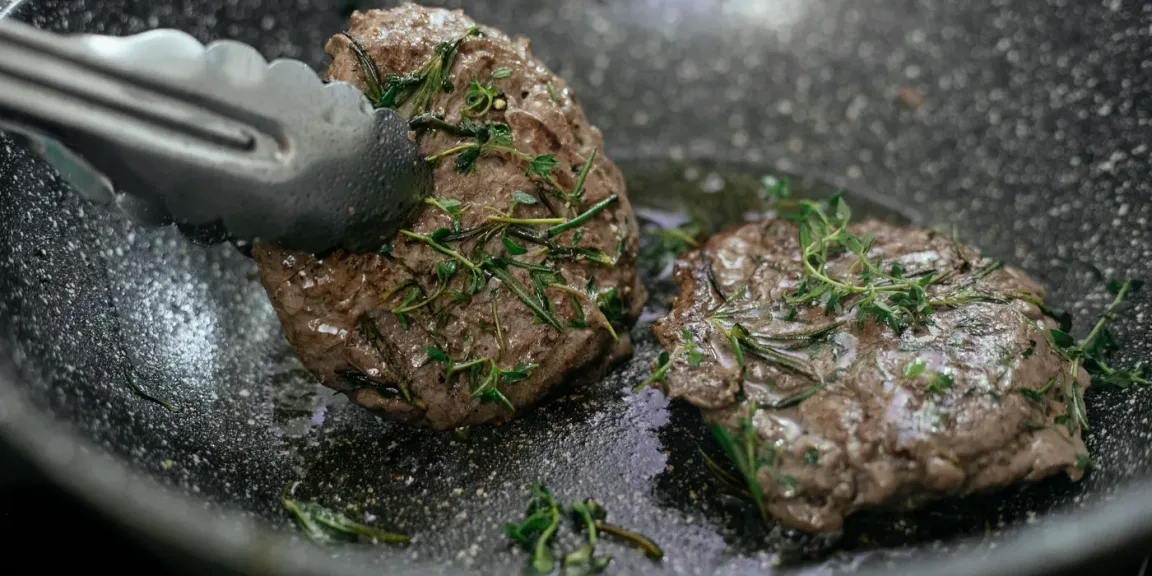 In einer schwarzen Pfanne braten zwei gewürzte Steaks mit Rosmarin und Thymian, während eines davon mit einer Küchenzange gewendet oder angehoben wird.