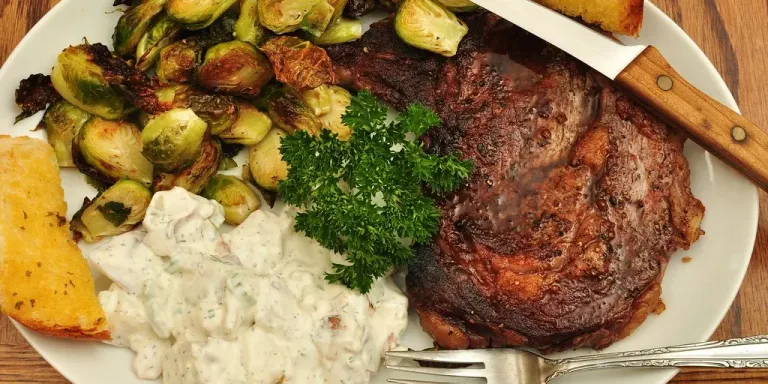 Auf einem weißen Teller liegen ein gebratenes Steak, gerösteter Rosenkohl, eine cremige Beilage, Petersilie und Brot; daneben liegen Messer und Gabel auf einem Holztisch.