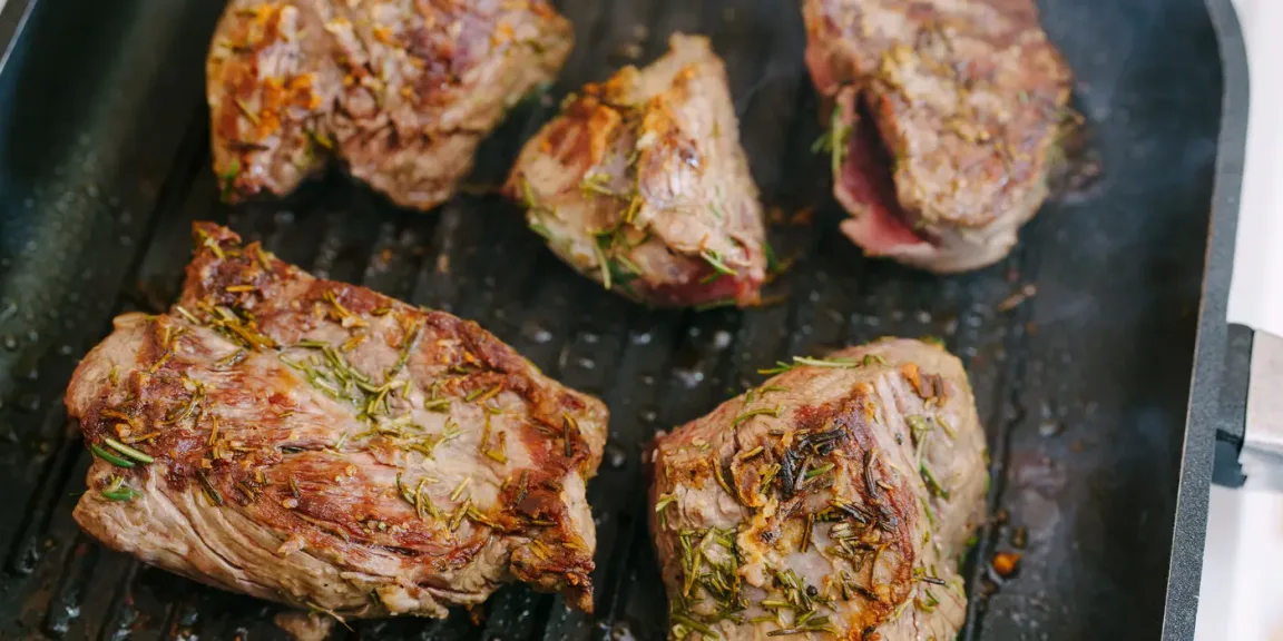 In einer geriffelten Grillpfanne liegen fünf mit Rosmarin und Gewürzen belegte, angebratene Steaks, von denen eines an der Seite noch rötliches Fleisch zeigt.