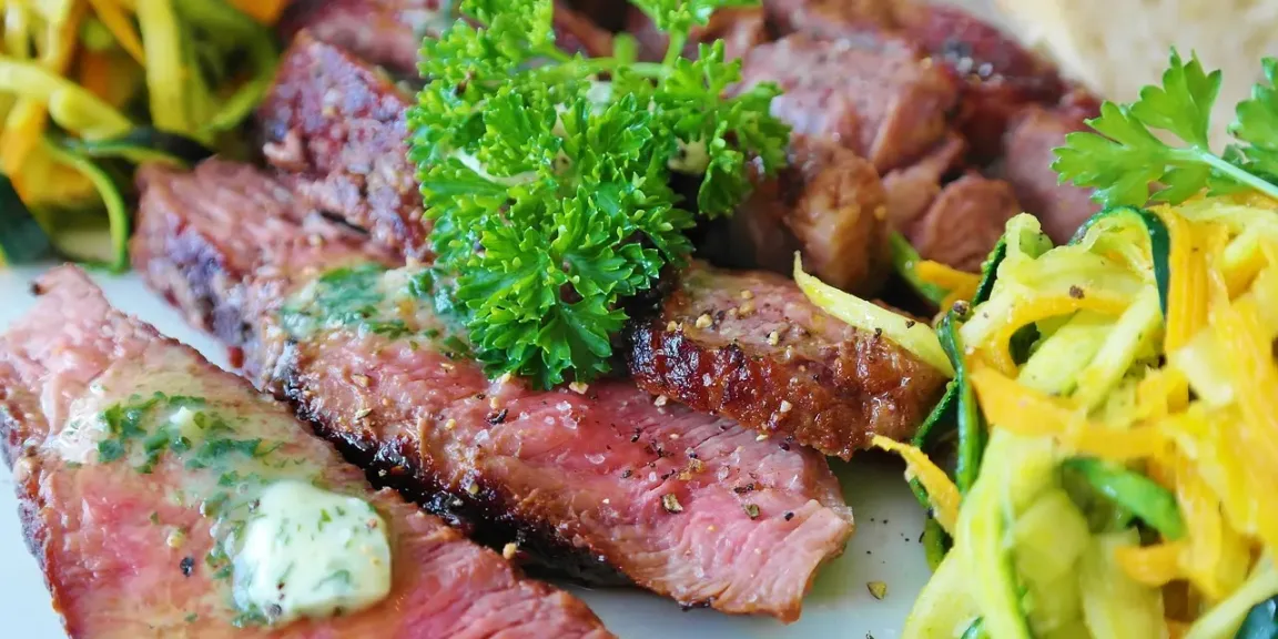 Auf einem Teller liegen mehrere aufgeschnittene, angebratene Steakstücke mit Kräuterbutter, Petersilie, Pfeffer und einer Beilage aus gelbem und grünem Gemüse.