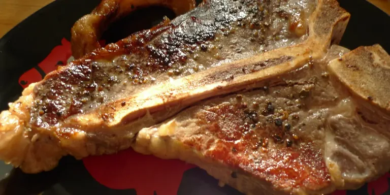 Auf einem schwarzen Teller liegen mehrere angebratene Steaks mit Knochen, gebräunter Kruste und sichtbaren Pfefferkörnern; im Hintergrund ist eine hölzerne Küchenoberfläche zu sehen.