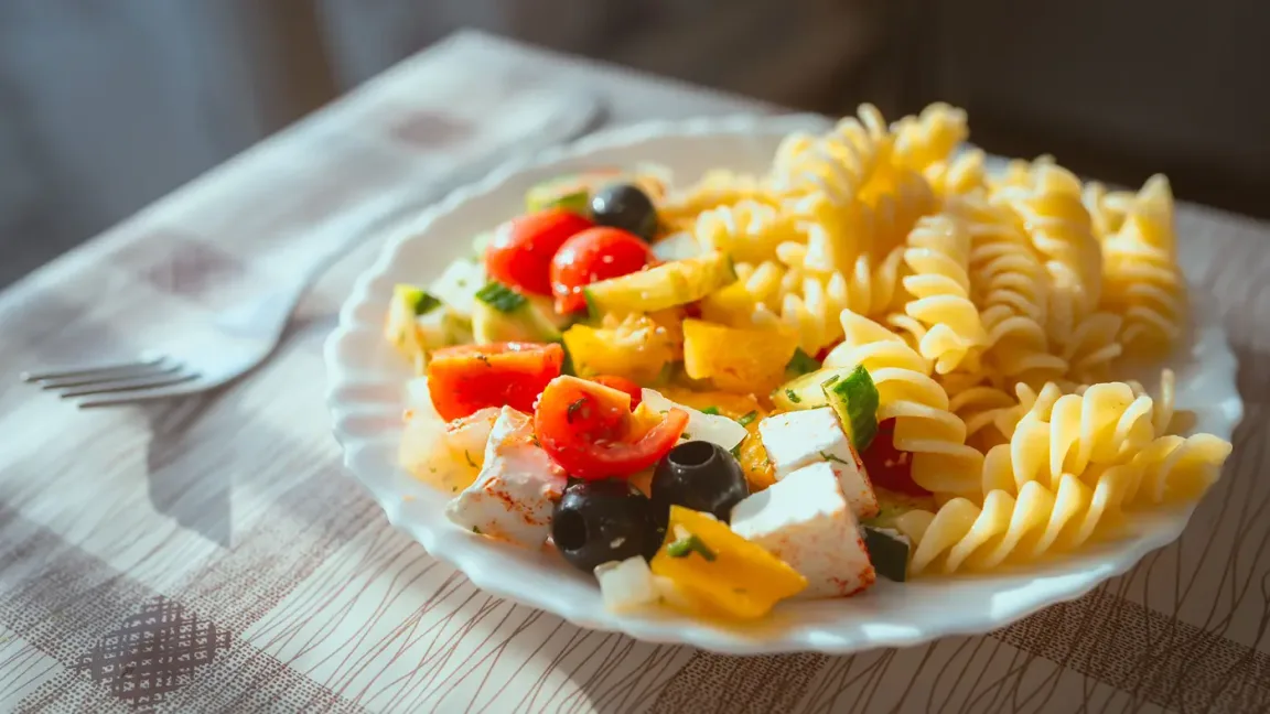 Auf einem weißen Teller liegt ein Nudelsalat aus Spiralnudeln, Tomaten, Paprika, Gurke, Oliven und Käse auf einem gedeckten Tisch neben einer Gabel.