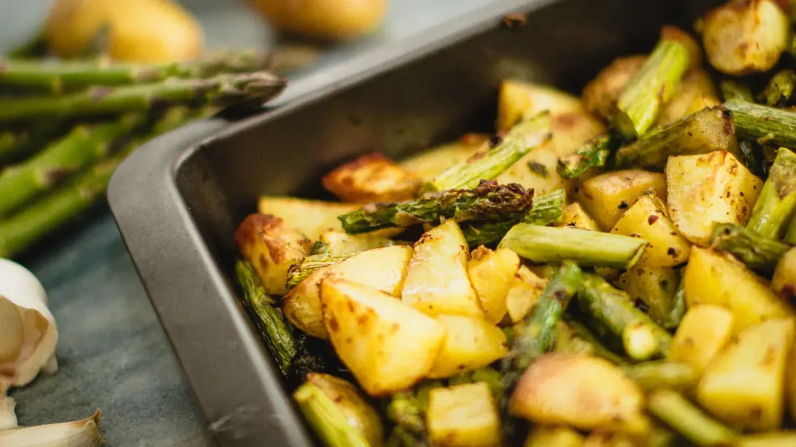 In einer dunklen Backform liegt geröstetes Ofengemüse aus Kartoffelstücken und grünem Spargel, daneben sind weiteres Gemüse und eine Knoblauchknolle auf einer Küchenarbeitsfläche zu sehen.