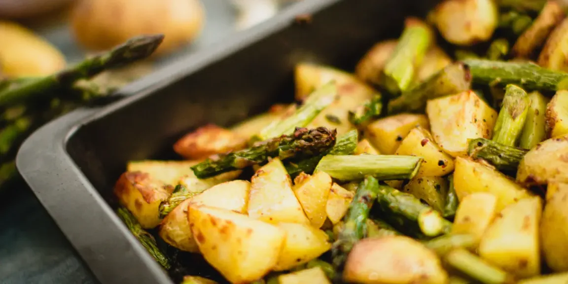 In einem dunklen Backblech liegt geröstetes Ofengemüse aus Kartoffelstücken, grünem Spargel und weiteren Gemüsestücken; im Hintergrund sind zusätzlich ungegarte Zutaten zu sehen.