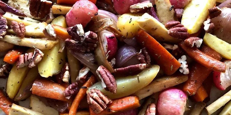 Zu sehen ist eine Nahaufnahme von verschiedenem gewürztem Ofengemüse wie Karotten, Kartoffeln, Pastinaken, Zwiebeln und Radieschen, das mit Pekannüssen in einer Schale oder auf einem Blech liegt.