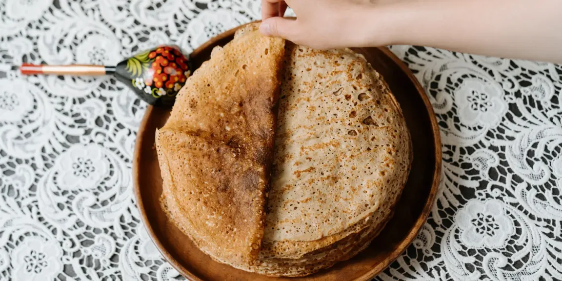Auf einem Holzteller liegen mehrere gefaltete Pfannkuchen, während eine Hand den obersten Pfannkuchen anhebt; daneben liegt ein dekorierter Pfannenwender auf einer weißen Spitzentischdecke.