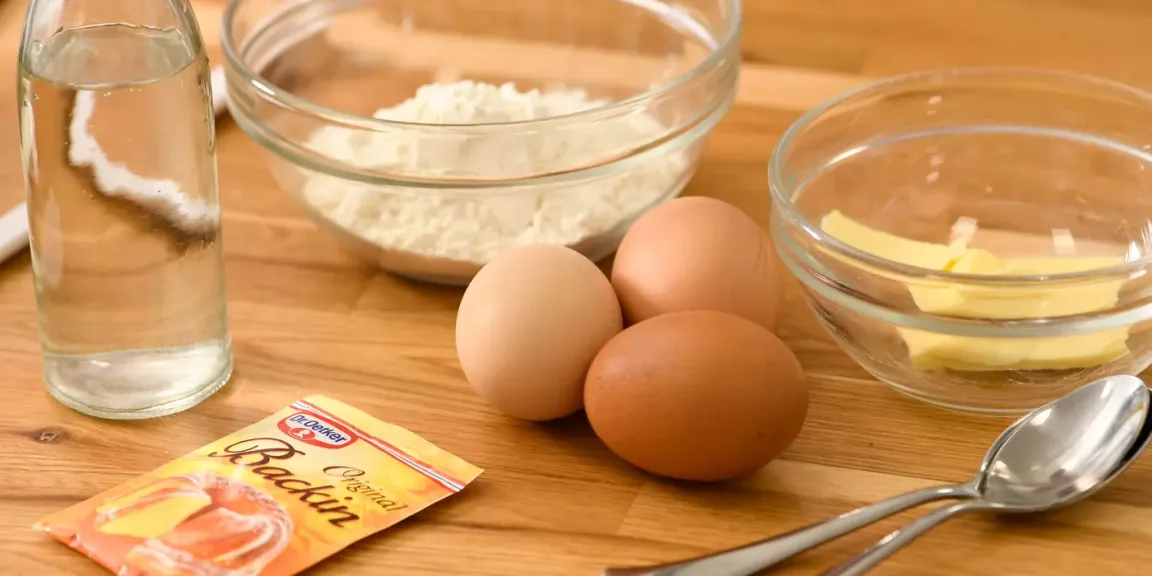 Auf einer hölzernen Küchenarbeitsfläche stehen eine Flasche mit Flüssigkeit, eine Schüssel Mehl, drei Eier, eine Glasschüssel mit Butter, zwei Löffel und eine Packung Backpulver.