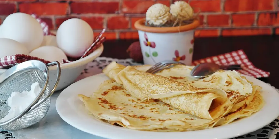 Auf einem weißen Teller liegen mehrere gefaltete, goldbraun gebackene Pfannkuchen; daneben stehen eine Schüssel mit Eiern, ein Sieb mit Mehl, ein Eisbecher und Besteck auf einem gedeckten Tisch vor einer Ziegelwand.