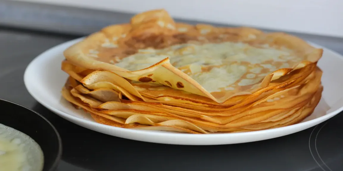 Auf einem weißen Teller liegt in einer Küche ein Stapel dünner, goldbraun gebackener Pfannkuchen auf einem schwarzen Kochfeld.