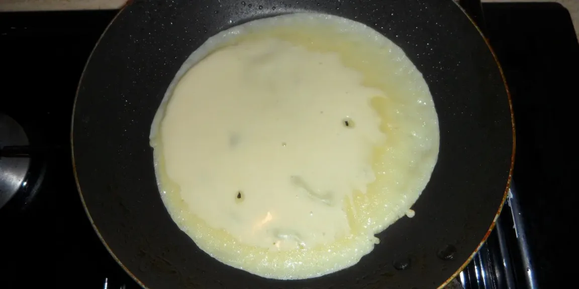 In einer beschichteten Pfanne auf einem Kochfeld liegt ein heller, noch nicht vollständig gebackener Pfannkuchen.