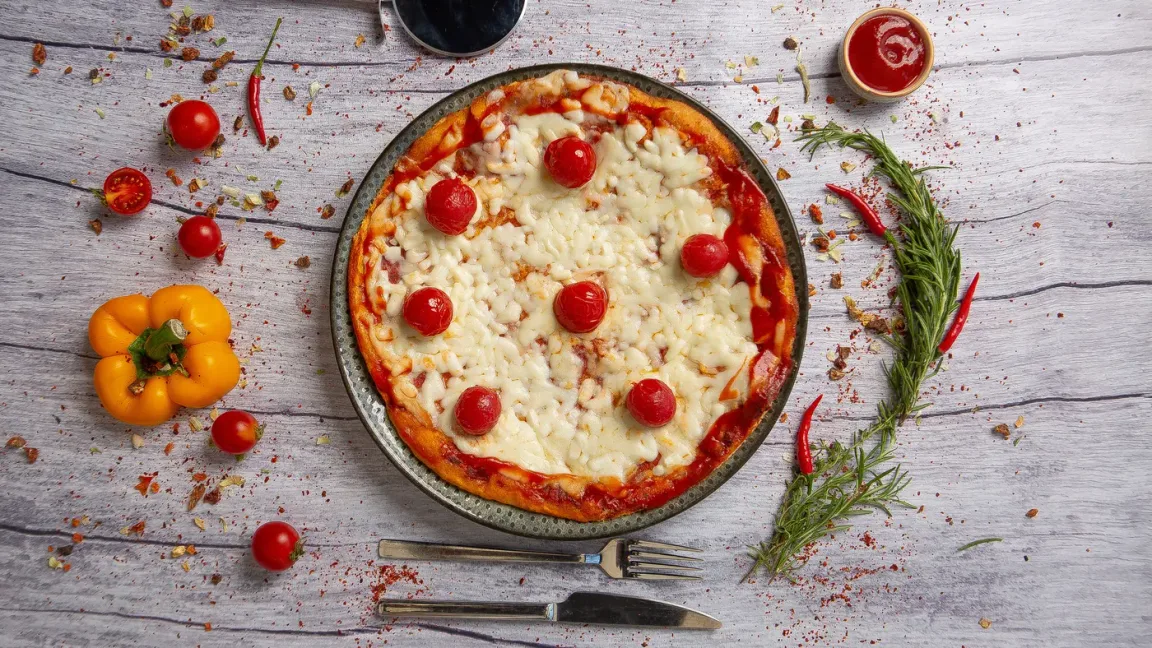 Auf einem Teller liegt eine runde, mit geschmolzenem Käse, Tomatensauce und Kirschtomaten belegte Pizza auf einem Holztisch, umgeben von Gemüse, Kräutern, Gewürzen sowie Messer und Gabel.