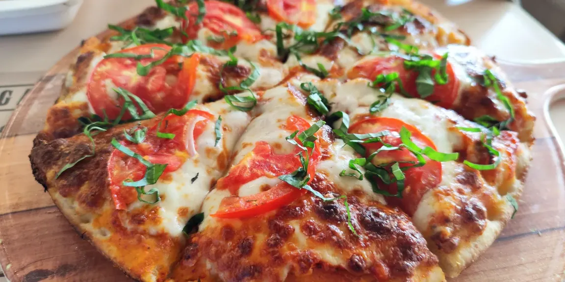 Eine belegte Pizza mit geschmolzenem und gebräuntem Käse, Tomatenscheiben und grünen Kräutern liegt auf einem Holzbrett in einer Küchenszene.