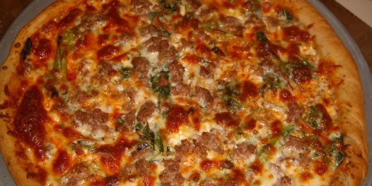 Eine große, gebackene Pizza mit goldbraunem Rand, geschmolzenem Käse, Tomatensauce sowie Fleischstücken und grünem Gemüse liegt auf einem runden Blech oder Teller.