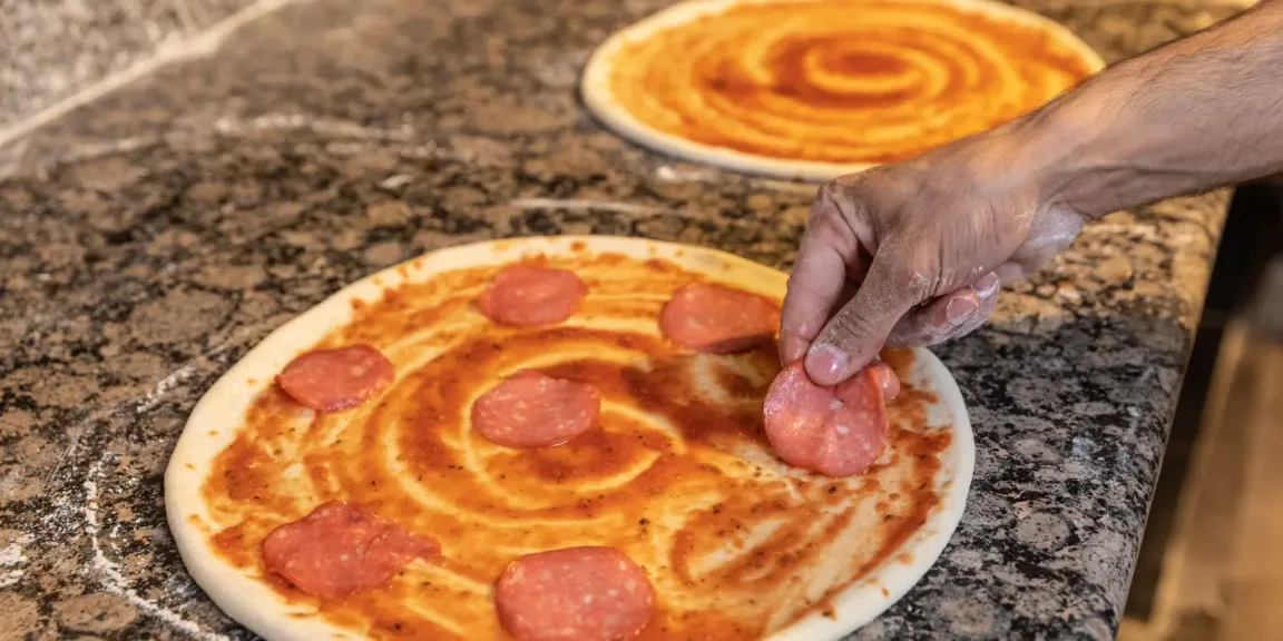 Auf einer Küchenarbeitsplatte bestreicht eine Hand einen runden Pizzateig mit Tomatensauce und belegt ihn mit Salamischeiben; im Hintergrund liegt ein weiterer mit Sauce bestrichener Teig.