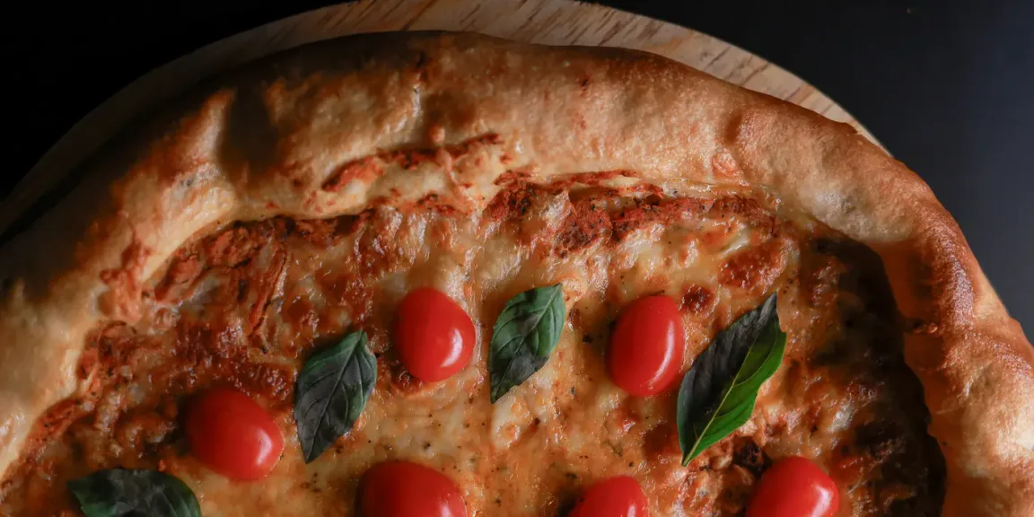 Eine gebackene Pizza mit gebräuntem Käse, Tomatensauce, Kirschtomaten und Basilikum liegt auf einem runden Holzbrett in einer dunklen Küchenumgebung.