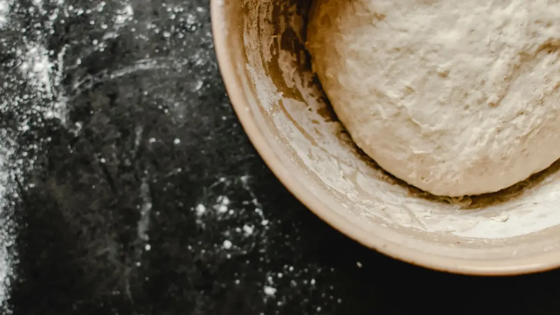 Auf einer bemehlten dunklen Arbeitsfläche steht rechts eine große Schüssel mit einem runden, bemehlten Teigballen darin.