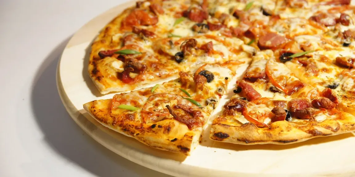 Auf einem hellen runden Teller liegt eine aufgeschnittene Pizza mit gebräuntem Teigboden, geschmolzenem Käse, Tomatensauce, Pilzen, Oliven, Paprika und Wurststücken.