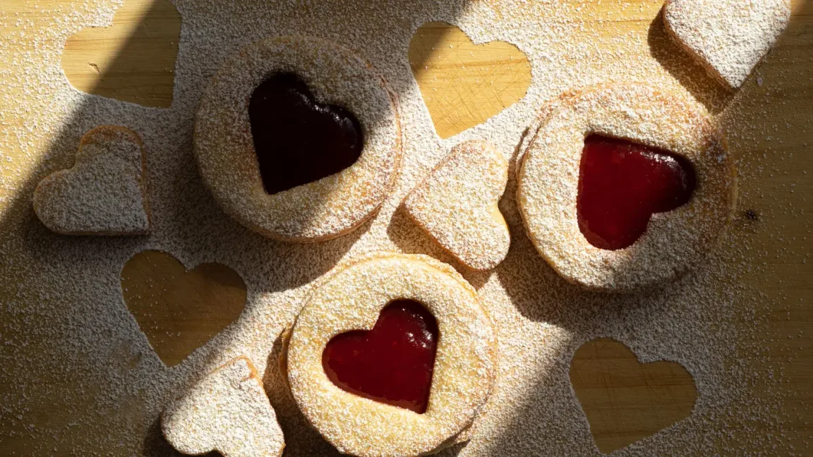 Auf einem bemehlten Holzbrett liegen mehrere herzförmige Plätzchen, teils mit herzförmiger Öffnung und roter Marmeladenfüllung, bestäubt mit Puderzucker.