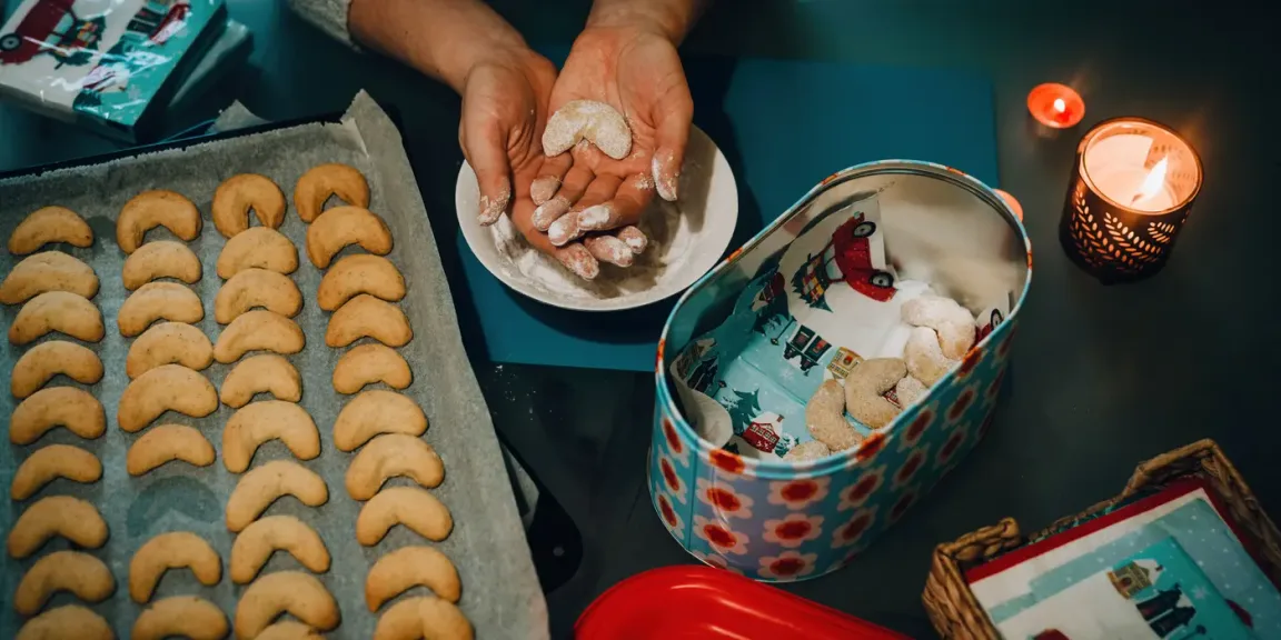 Eine Person hält geformte, mit Puderzucker bestäubte halbmondförmige Plätzchen über einer Schale; daneben liegen weitere gebackene Plätzchen auf einem Backblech und in einer dekorierten Keksdose.
