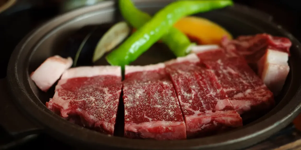 In einer dunklen Schmorpfanne liegen mehrere dicke Stücke rohes, gewürztes Rindfleisch mit sichtbarer Marmorierung, dazu grüne Paprika, Zwiebel und weiteres Gemüse.