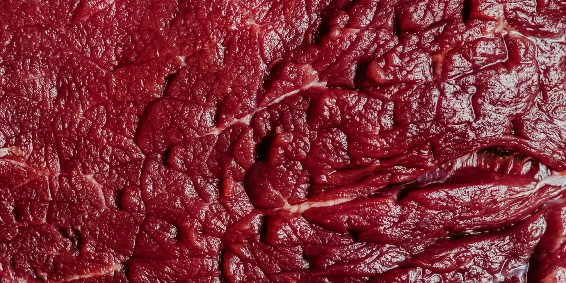 Eine Nahaufnahme zeigt mehrere rohe, dunkelrote Stücke Rindfleisch mit sichtbaren Muskelfasern, Fett und Bindegewebe auf einer nicht erkennbaren Unterlage.