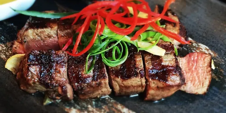 Auf einem dunklen Teller liegen mehrere Scheiben gebratenes Rindfleisch mit sichtbaren Grillstreifen, garniert mit roten und grünen Gemüsestreifen.