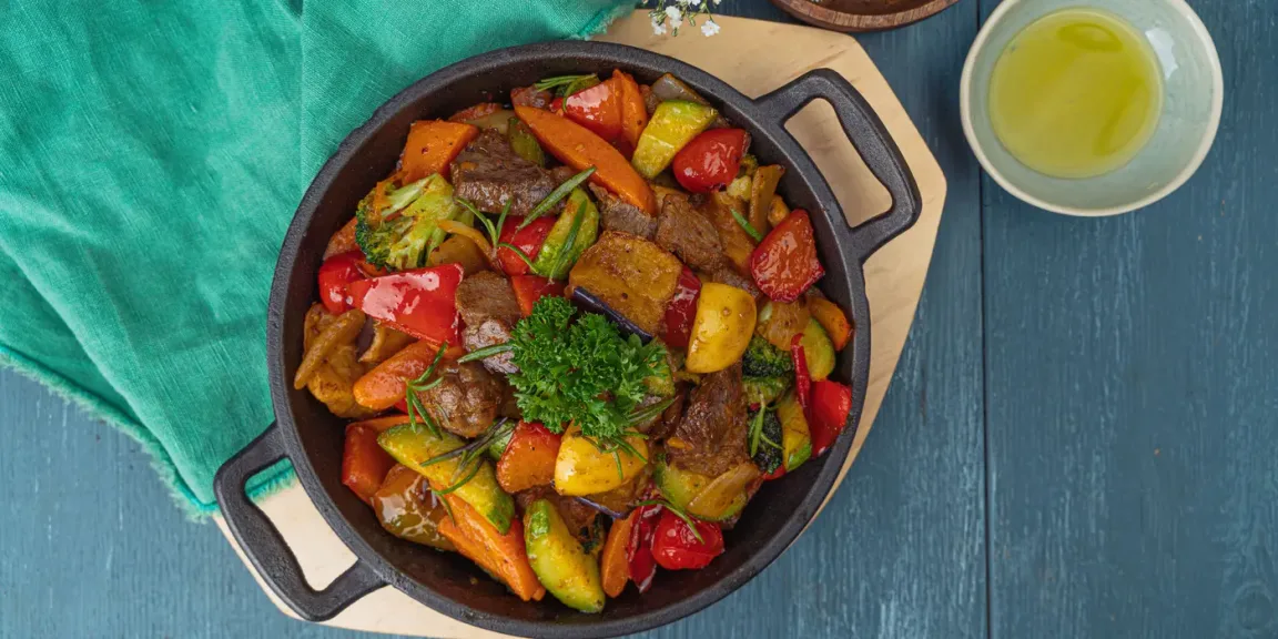Zu sehen ist eine schwarze gusseiserne Pfanne mit gebratenen Rindfleischstücken, Kartoffeln und verschiedenem Gemüse auf einem Holzbrett, daneben steht eine Schale mit heller Flüssigkeit.
