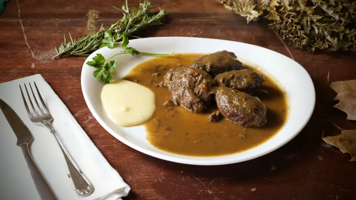Auf einem weißen Teller liegen mehrere geschmorte Rinderrouladen in brauner Soße neben einem Klecks Kartoffelpüree, garniert mit Petersilie und Kräutern, auf einem Holztisch mit Besteck.