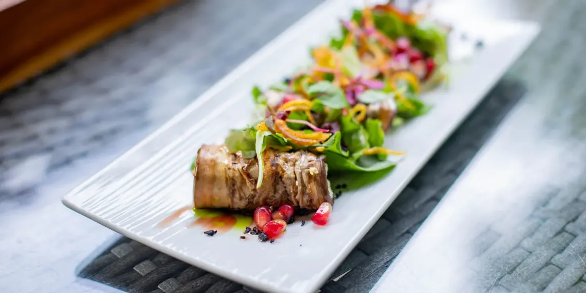 Auf einem länglichen weißen Teller liegt eine aufgeschnittene gefüllte Rinderroulade neben einem bunten Blattsalat mit Gemüse und Granatapfelkernen auf einem Tisch.