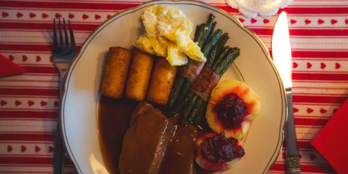 Auf einem Teller liegen zwei mit dunkler Soße bedeckte Rinderrouladen neben Kroketten, grünen Bohnen, Kartoffelklößen mit roter Beilage und einer Portion Kartoffelsalat; der Teller steht auf einer rot-weiß gemusterten Tischdecke mit Besteck daneben.