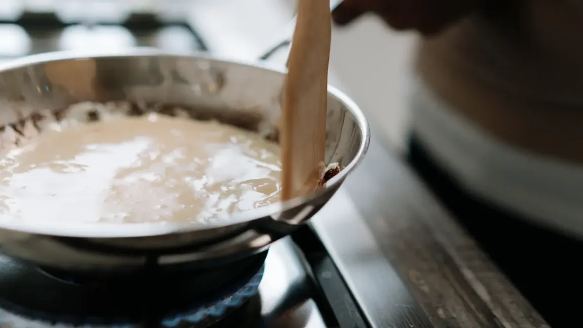 Helle Soße wird in einer Edelstahlpfanne auf dem Gasherd mit dem Holzlöffel gerührt