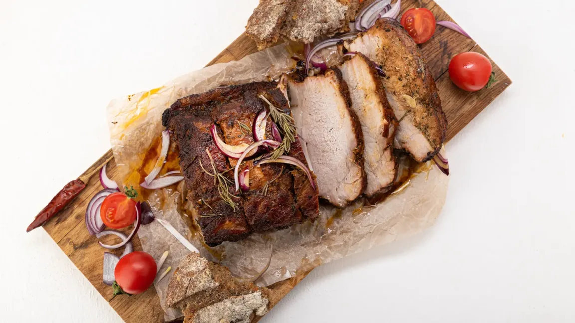 Auf einem mit Backpapier belegten Holzbrett liegt ein gebratener Schweinebraten, teils im Ganzen und teils in Scheiben geschnitten, garniert mit roten Zwiebeln, Rosmarin und Tomaten.
