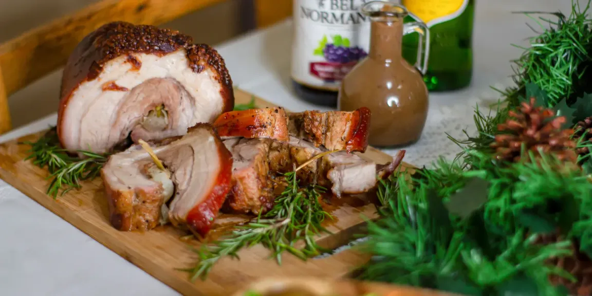 Auf einem Holzbrett liegen ein aufgeschnittener, gerollter Schweinebraten und mehrere Scheiben davon, dekoriert mit Rosmarinzweigen; im Hintergrund stehen Küchengefäße und Flaschen.