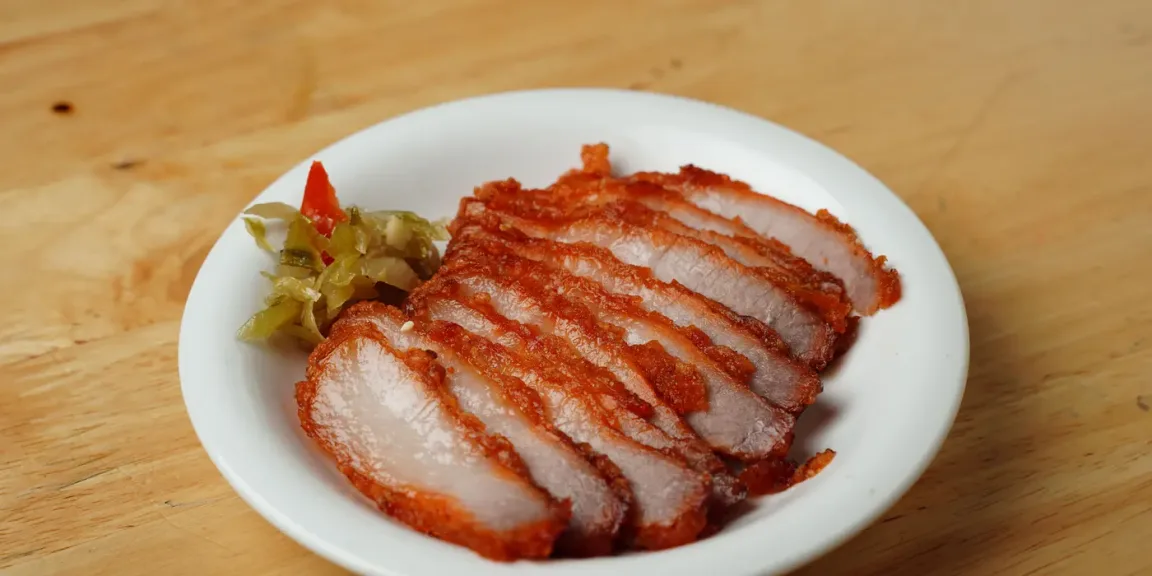Auf einem weißen Teller liegen mehrere Scheiben knusprig gebratenen Schweinebratens neben etwas eingelegtem Gemüse auf einem Holztisch.