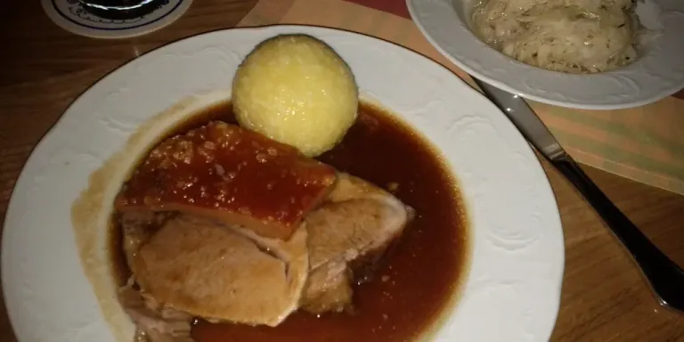 Auf einem weißen Teller liegen mehrere Scheiben Schweinebraten mit knuspriger Kruste in dunkler Bratensoße, dazu ein Kartoffelknödel; daneben steht ein Teller mit Kraut auf einem gedeckten Holztisch.