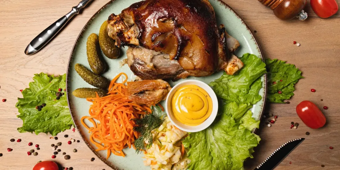 Auf einem Teller liegt ein gebräunter Schweinebraten mit knuspriger Kruste, dazu sind Gewürzgurken, Salat, geraspelte Möhren, Krautsalat und eine Schale Senf auf einem Holztisch angerichtet.