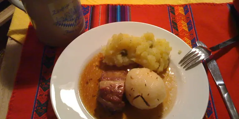 Auf einem weißen Teller liegen mehrere Scheiben Schweinebraten in brauner Soße neben einer Kartoffel und einer Portion Kartoffelpüree; daneben stehen Besteck und ein bedruckter Becher auf einem bunten Tischset.