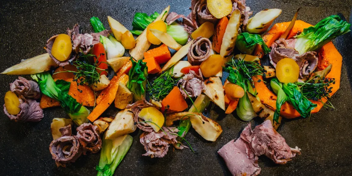 Auf einer dunklen Platte liegen aufgeschnittene Stücke gegartes Rindfleisch zusammen mit Möhren, Pastinaken oder anderem Wurzelgemüse, grünem Blattgemüse und Kräutern.