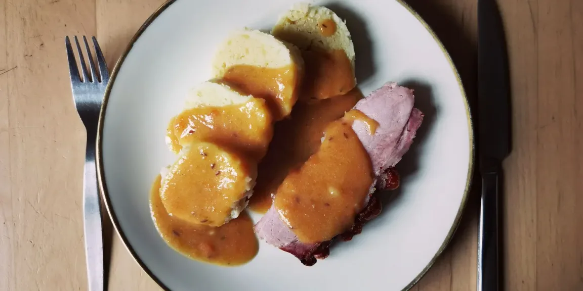 Auf einem weißen Teller liegen mehrere Scheiben Kartoffelklöße und aufgeschnittenes rosa gegartes Fleisch, beides mit brauner Soße übergossen, daneben liegen Messer und Gabel auf einem Holztisch.