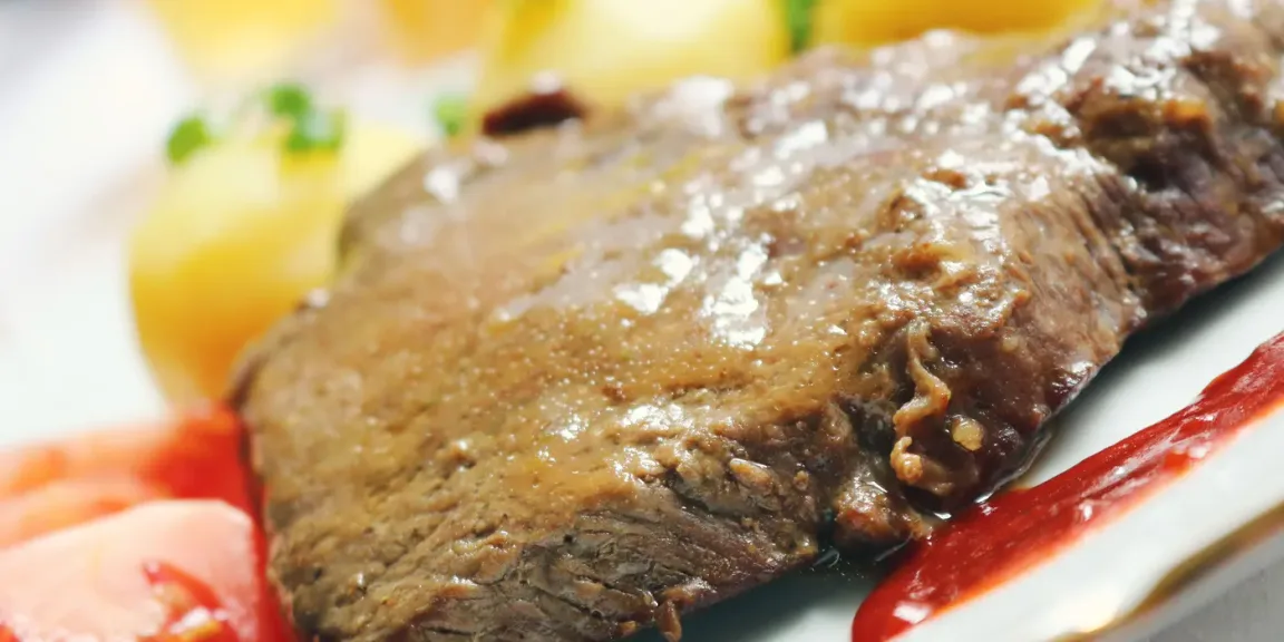 Auf einem weißen Teller liegt ein gebräuntes, längliches Stück gegartes Rindfleisch, umgeben von Kartoffeln, Gemüse und Kräutern.
