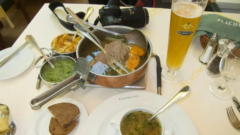 Auf einem gedeckten Restauranttisch stehen eine Kupferpfanne mit gekochtem Rindfleisch in Brühe und Gemüse, Schalen mit Beilagen, Brot und Suppe sowie ein Bierglas und Besteck; im Hintergrund liegt eine Kamera.