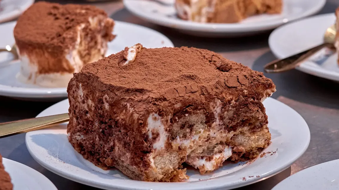Auf einem weißen Teller liegt ein angeschnittenes, mit Kakaopulver bestäubtes Stück Tiramisu mit sichtbaren Lagen aus getränkten Biskuitstücken und heller Creme; im Hintergrund stehen weitere Teller mit Tiramisu und Besteck auf einem Tisch.