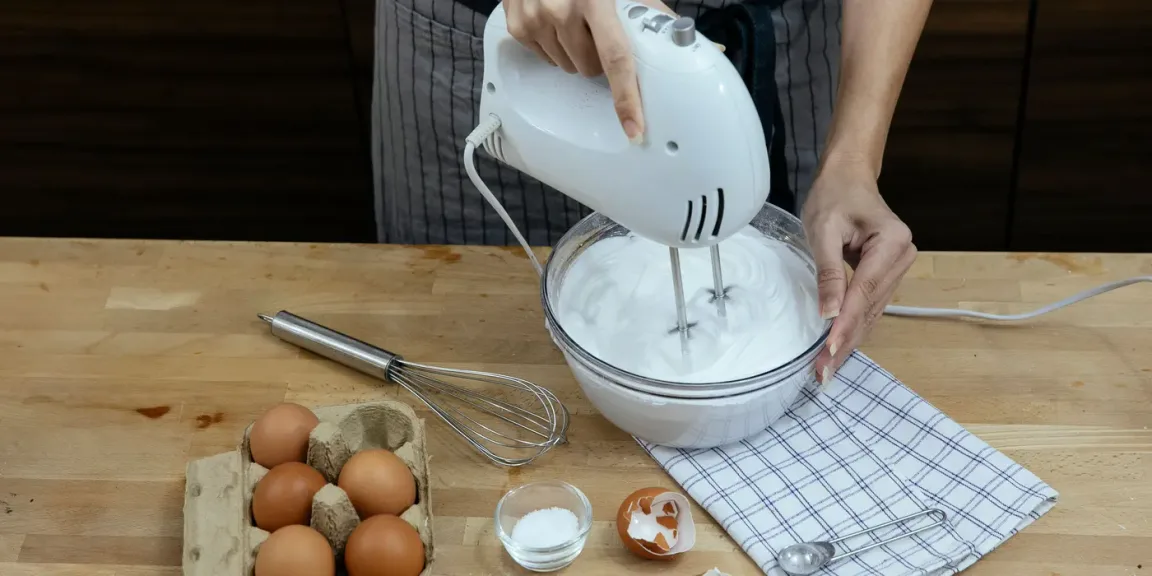 Eine Person schlägt mit einem elektrischen Handmixer eine weiße Masse in einer Glasschüssel auf einer hölzernen Arbeitsfläche, daneben liegen Eier, ein Schneebesen, eine Schale mit weißem Pulver, Eierschalen und ein Küchentuch.