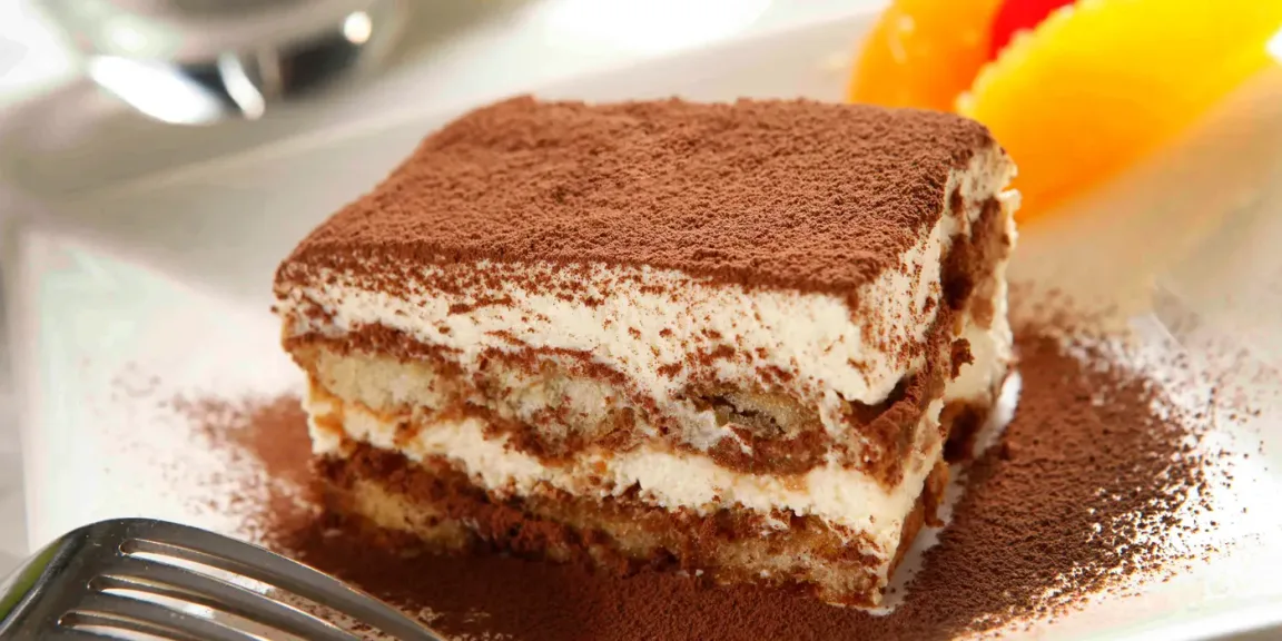 Auf einem weißen Teller liegt ein rechteckiges Stück Tiramisu mit mehreren Schichten aus kakaobestäubter Mascarponecreme und getränktem Biskuit; daneben liegen eine Gabel und unscharf sichtbare Orangenspalten.