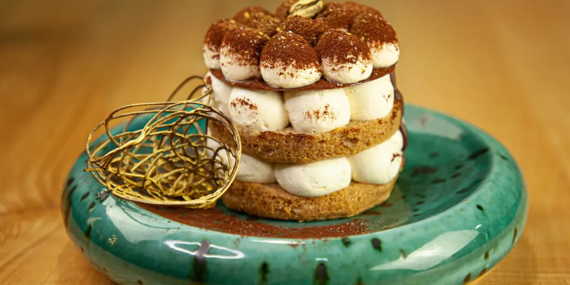 Auf einem türkisfarbenen Teller steht ein geschichtetes Tiramisu mit Biskuitböden, weißer Creme und Kakaopulver, daneben liegt ein dekoratives goldenes Körbchen auf einer Holzoberfläche.