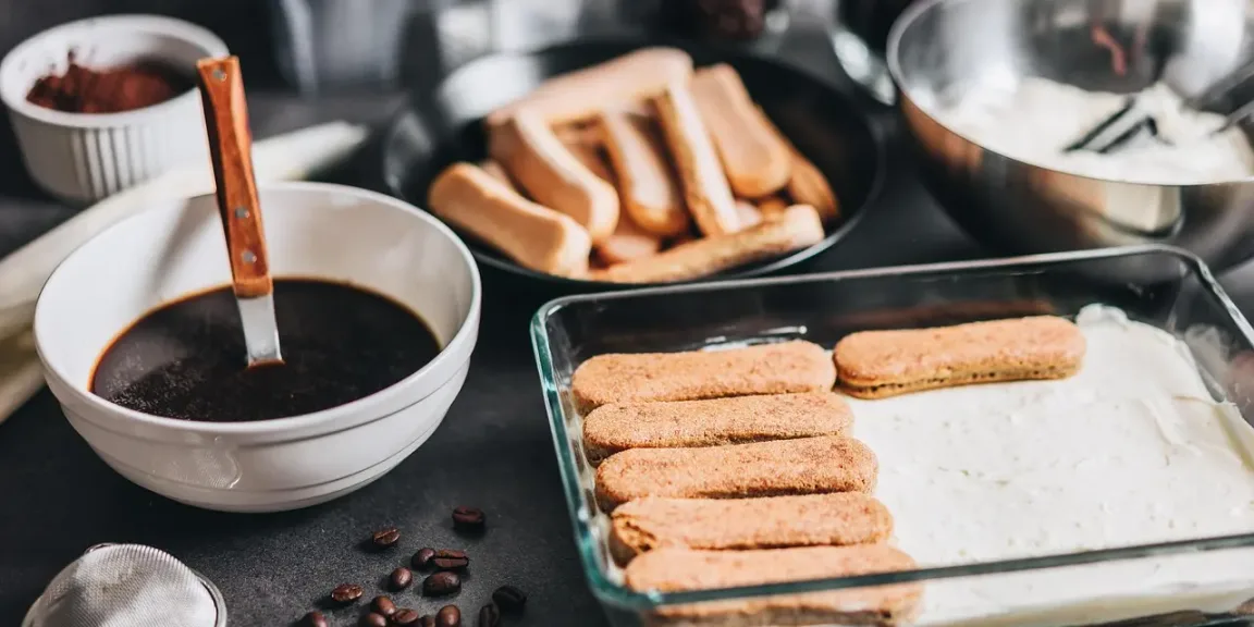 Auf einer Küchenarbeitsfläche stehen eine Schale mit dunklem Kaffee und Löffel, eine Schale mit weißer Creme sowie Teller mit Löffelbiskuits; in einer rechteckigen Glasschale wird Tiramisu aus Biskuits und Creme geschichtet.
